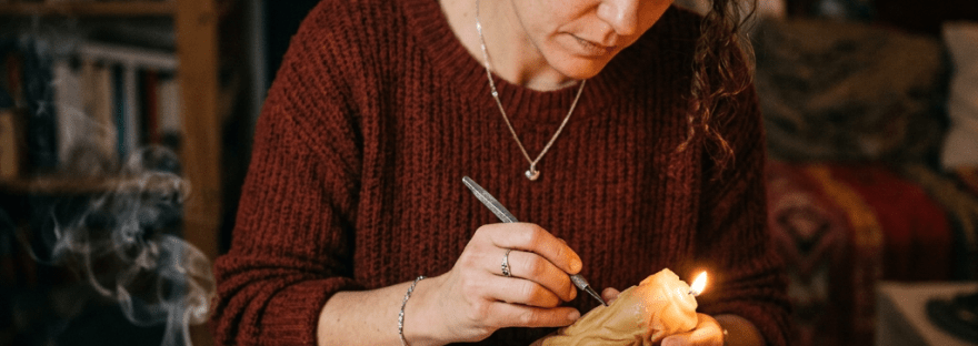 Woman carving a lit ritual candle at a table with crystals, dried flowers, and handwritten notes
